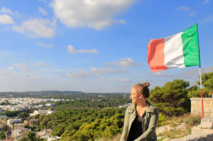 woman sits in front of a lookout in Italy with the italian flag behind her